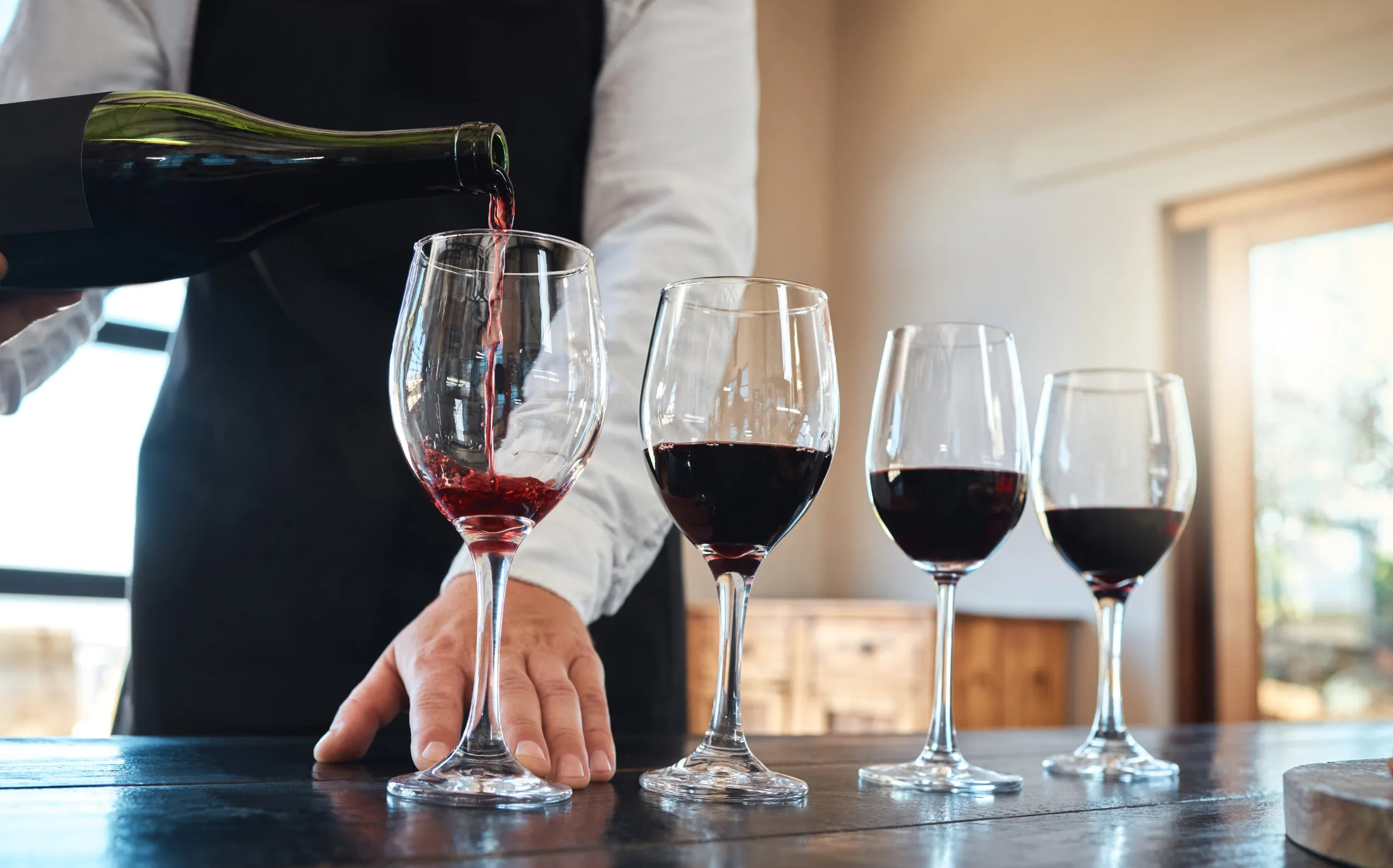 A person in an apron pouring red wine into the first of four glasses