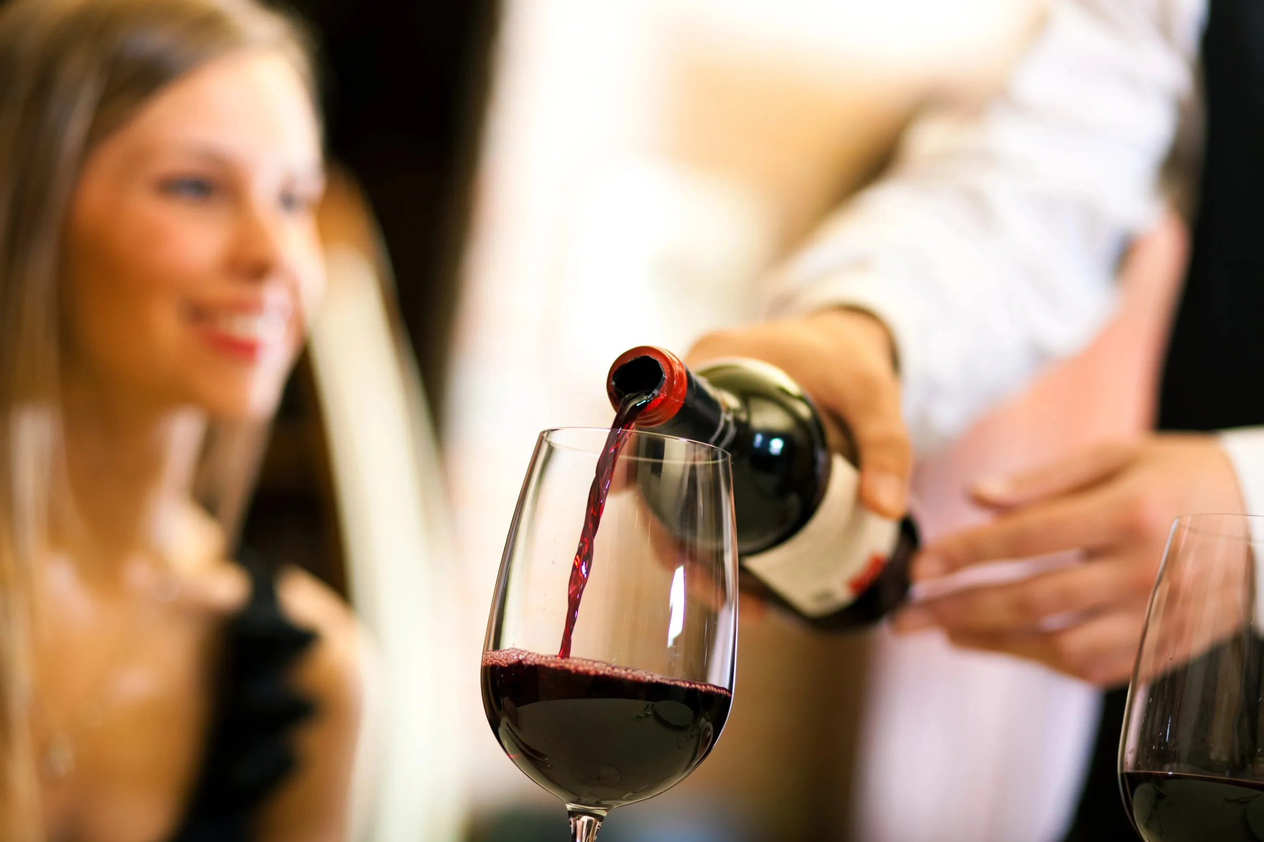 Red wine pouring from a bottle into a glass, with a guest out of focus in the background