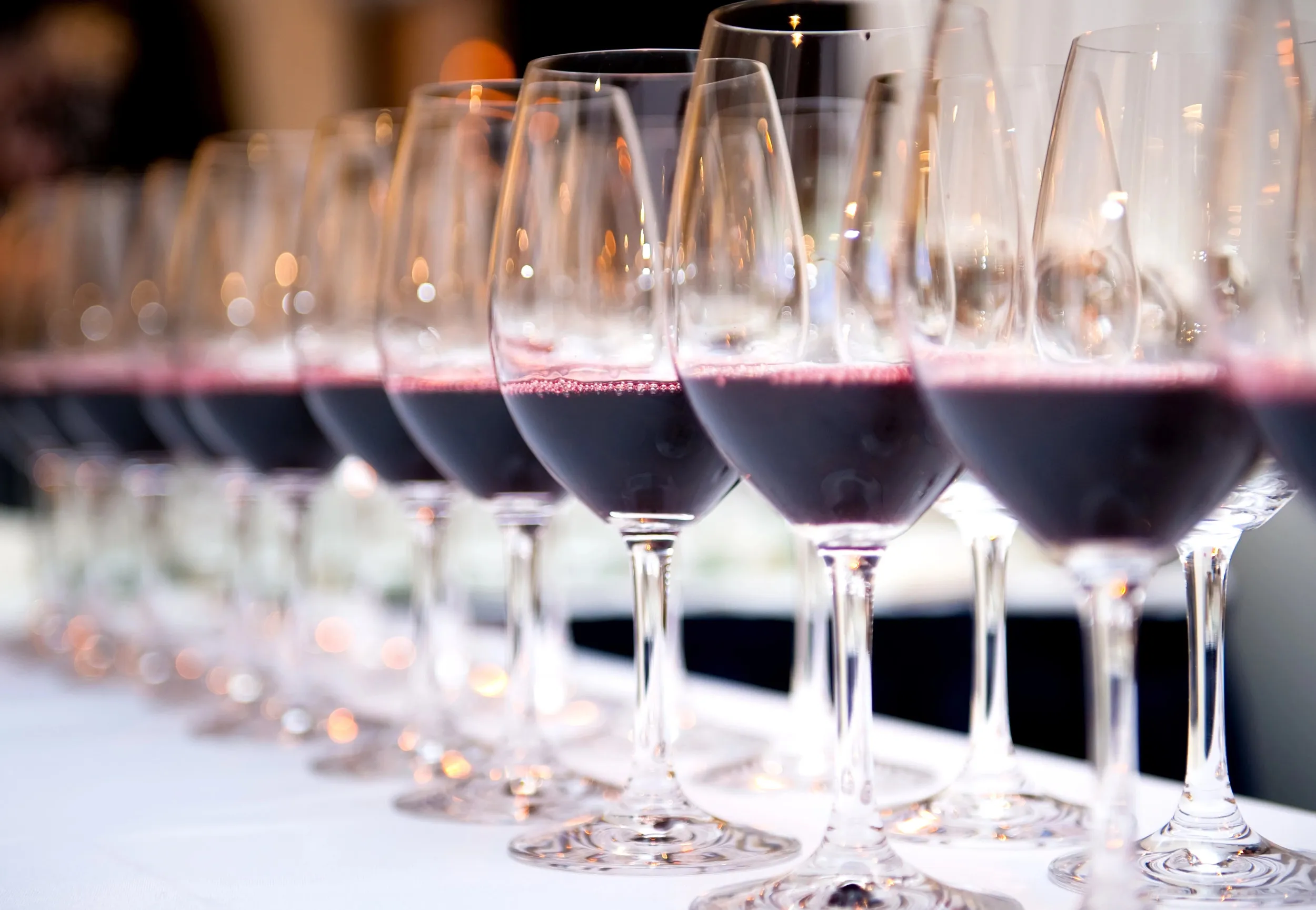 A diagonal row of stemmed glasses, each with a small pour of red wine